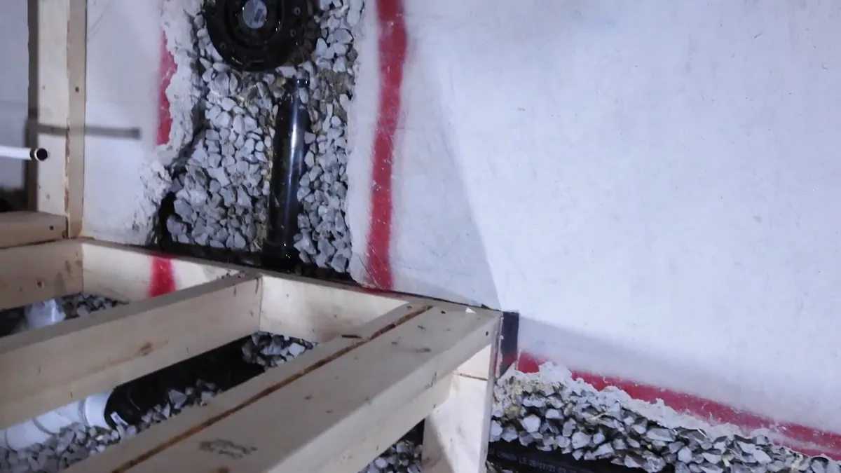 Black drain pipe bedded in gravel inside a framed floor section, with a white pipe beside it